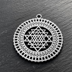 May include: A silver-colored pendant with a geometric design. The pendant features a central Sri Yantra symbol composed of interlocking triangles, surrounded by a decorative border. It has a small loop at the top for hanging.