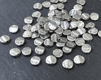 May include: A collection of small, round, silver-colored metal beads with a hammered, textured surface. Each bead features a slightly irregular, flattened shape and a single hole drilled through the center for threading, scattered across the frame.