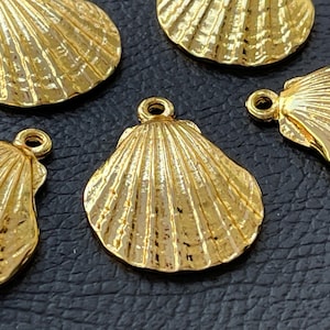 May include: Close-up of several gold-colored seashell charms. Each charm has a detailed, ridged texture and a small loop at the top for attaching to jewelry. The charms are arranged on a dark surface.