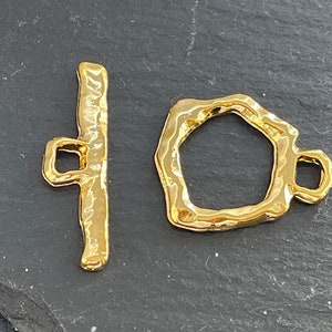 May include: A gold-coloured toggle clasp set. The clasp features a textured bar and an irregular-shaped ring with a small loop. The metal has a hammered appearance, giving it a handcrafted look. Suitable for jewellery making.