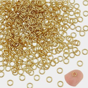 Könnte beinhalten: Ein großer Haufen goldener Sprungringe, ein gängiges Material für die Schmuckherstellung. Die Ringe sind auf einem weißen Hintergrund verstreut. Eine Nahaufnahme eines einzelnen Sprungrings wird auf einem Finger gezeigt.