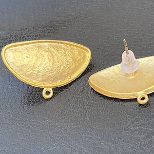 May include: A pair of gold-toned earrings with a half-circle shape. The earrings have a textured surface and a small loop on the bottom.