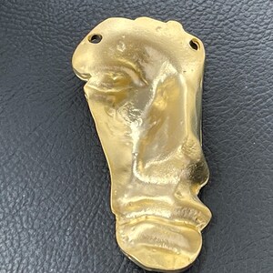 Könnte beinhalten: Ein goldfarbener Metallanhänger in Form eines menschlichen Fußabdrucks. Die Oberfläche hat ein glattes, reflektierendes Finish mit organischen, unebenen Konturen, die die Textur einer Fußsohle nachahmen. Zwei kleine Löcher befinden sich oben.
