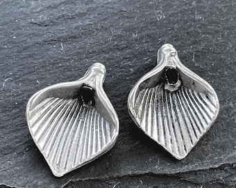 May include: Two silver-toned metal charms shaped like calla lily flowers. Each charm features a flared, petal-like form with deep, vertical ridges radiating from a central dark opening at the top, creating a textured, metallic finish.