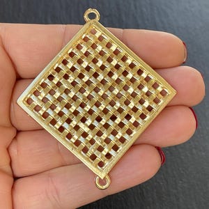 May include: A gold-colored, diamond-shaped jewelry component with a woven pattern. The component has a square grid design within a diamond frame. It features a small loop at the top and bottom for attachment. The piece is held in a hand.