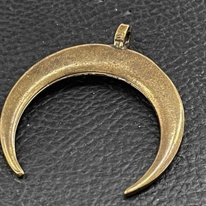 Könnte beinhalten: Ein goldfarbener Halbmond-Anhänger mit einer Öse oben zum Anbringen an eine Halskette oder ein Armband.