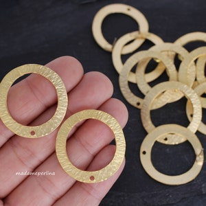 Può includere: Primo piano di due cerchi in metallo dorato con una texture martellata. I cerchi hanno un foro al centro e misurano circa 5 cm di diametro. I cerchi sono su una mano e ci sono molti altri cerchi sullo sfondo.