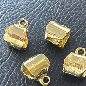 May include: Four small, gold-colored metal jewelry findings with a polished, reflective surface. Each piece features a rounded, irregular shape with a small circular loop attached at the top, designed for connecting components in jewelry making.