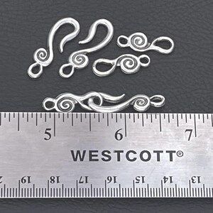 May include: A set of silver-toned metal jewelry clasps featuring a spiral design. The clasps include hook and loop components, with one pair connected. A Westcott ruler is positioned below the items, showing a length of approximately 1.75 inches.