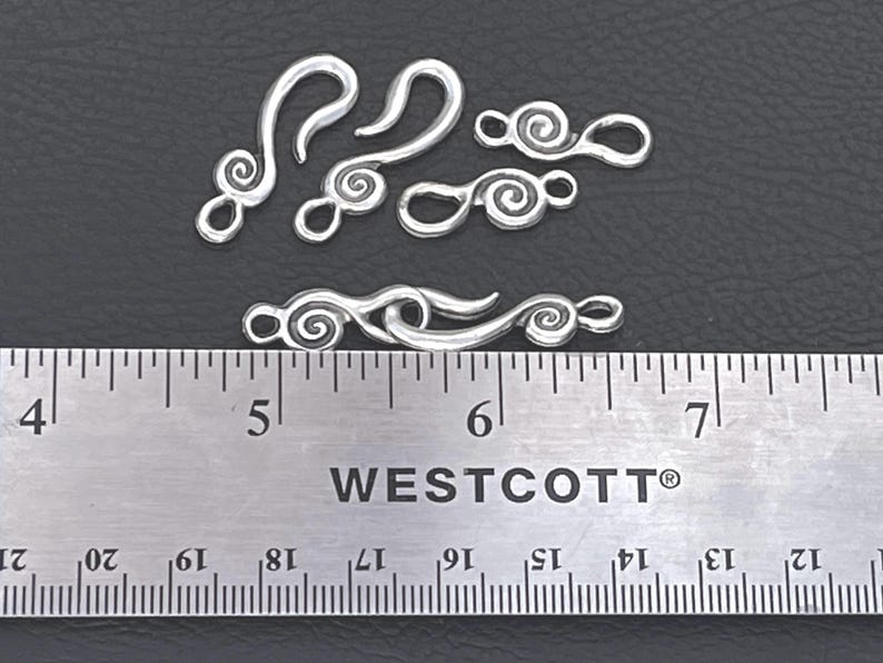 May include: A set of silver-toned metal jewelry clasps featuring a spiral design. The clasps include hook and loop components, with one pair connected. A Westcott ruler is positioned below the items, showing a length of approximately 1.75 inches.