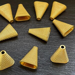 May include: A collection of ten gold-colored, cone-shaped jewelry findings. Each finding has a textured, dotted surface and a wide, oval opening at the base. The findings are arranged on a dark surface, showcasing their metallic sheen and intricate design.