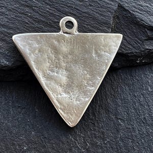 Op de afbeelding: Een zilveren driehoekige hanger met een glad, getextureerd oppervlak. De hanger heeft een lus aan de bovenkant om aan een ketting te bevestigen.
