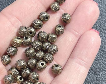 May include: A collection of small, round, metallic-toned beads with a textured, grid-like surface pattern. Each bead features a central hole for threading, displaying a dark, antique-style finish with raised bumps across the exterior surface.