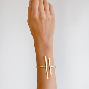 Op de afbeelding: Een gouden armband met een geometrisch ontwerp. De armband is gemaakt van drie dunne, rechte metalen staven die aan de uiteinden met elkaar verbonden zijn. De staven zijn parallel gerangschikt, waarbij de twee buitenste staven iets langer zijn dan de middelste staaf.