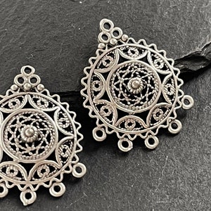 May include: Two silver-colored filigree pendants. Each pendant features a central spiral design surrounded by intricate openwork patterns. The pendants have a circular shape with a scalloped edge and small loops for attachment, perfect for jewelry making.