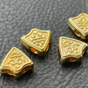 May include: Four gold-colored metal beads with intricate designs. Each bead has a triangular shape with rounded edges and two holes for stringing.