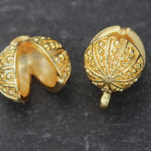 May include: Two gold-coloured jewellery components. One is open, revealing the interior, and the other is closed. Both have a textured, dotted surface and a small loop at the base. The components are on a dark grey surface.