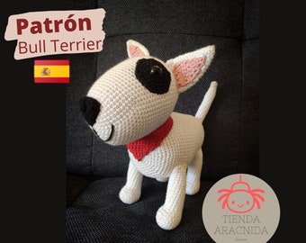May include: A white crocheted Bull Terrier amigurumi toy with black eyes and a red collar. The toy is standing on a gray couch. The image also includes the text "Patrón Bull Terrier" and a Spanish flag.