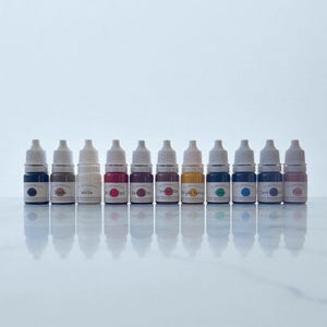 May include: A collection of ten small, clear bottles filled with colorful liquid, arranged in a row. Each bottle has a white cap and a label with text. The colors include shades of blue, brown, white, red, purple, yellow, and pink. The bottles are on a white surface.