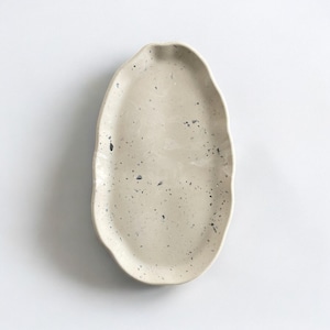 May include: A speckled, off-white ceramic serving platter with an organic, oval shape. The platter has a slightly textured surface and is dotted with small, dark gray specks. The edges are gently undulating, adding to its handcrafted appearance.