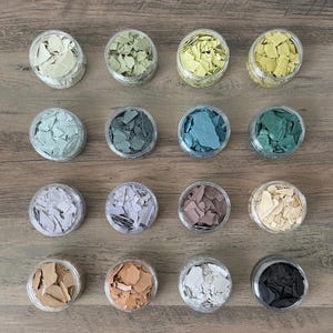 May include: Overhead shot of sixteen small, clear plastic jars filled with colorful, irregularly shaped paint chips. The chips are in various shades of green, blue, brown, and neutral tones, arranged in a grid pattern on a wooden surface.