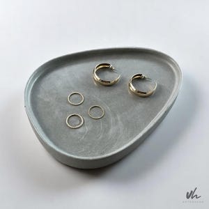 May include: A grey concrete tray with a rounded shape, featuring two gold hoop earrings and four gold rings. The tray is ideal for organising jewellery or other small items.