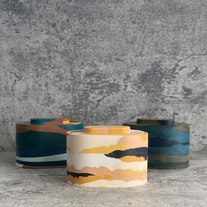 May include: Three oval-shaped containers with lids, each featuring a unique marbled pattern. The front container is cream with yellow, black, and tan swirls. The left container is blue, brown, and tan, while the right container is dark green and blue.