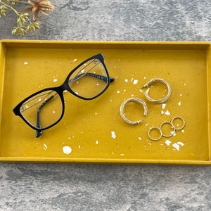 May include: A yellow tray with white flecks, featuring a pair of black eyeglasses, two gold hoop earrings, and three smaller gold rings.