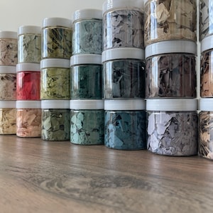May include: A row of small glass jars filled with colorful, iridescent flakes. The jars are arranged in a line on a wooden surface.