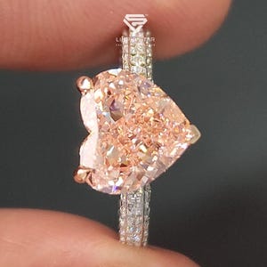 May include: A heart-shaped, peach-colored gemstone ring. The ring features a large, heart-shaped stone set on a band with smaller, clear stones. The ring is held between fingers against a dark background.