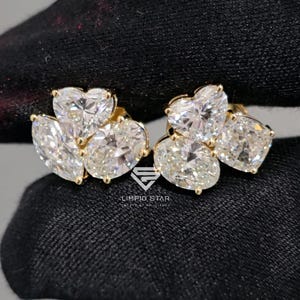 May include: A pair of gold stud earrings featuring multiple clear, sparkling gemstones in heart and oval shapes. The earrings are set against a black background, showcasing their brilliance.