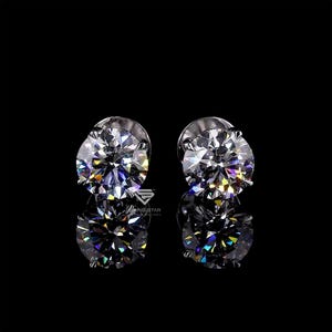 May include: A pair of round, brilliant-cut diamond stud earrings. The diamonds are set in a silver or white gold setting and reflect light with a rainbow of colors. The earrings are displayed on a reflective black surface.