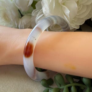 May include: A translucent bangle bracelet with a mix of white, gray, and reddish-brown hues. The bracelet is smooth and circular, with a prominent reddish-brown spot. The jewelry is worn on the wrist.