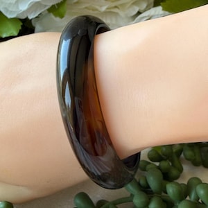 May include: A dark brown, glossy bangle bracelet. The smooth, rounded bracelet is worn on a wrist, showcasing its rich color and reflective surface. The jewelry piece is a classic accessory.
