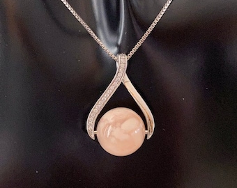 May include: A silver-toned pendant necklace featuring a large, round, peach-colored stone. The pendant has a curved, open design, embellished with small, clear crystals. The necklace chain is a delicate, square-link style.