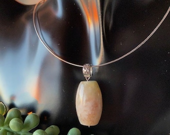 May include: A silver necklace with a pendant. The pendant is a light beige oval shape with a silver bail. The necklace is displayed on a black surface with green plant accents.