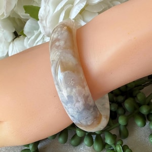 May include: A translucent, marbled bangle bracelet in shades of cream, tan, and gray. The smooth, polished surface reflects light, showcasing the natural patterns of the stone. The bracelet is a classic, round design, suitable for everyday wear.