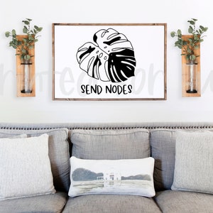 May include: A white framed print with a black and white illustration of a monstera leaf. The text "SEND NODES" is below the leaf. The print is hanging on a white wall above a gray couch with pillows.