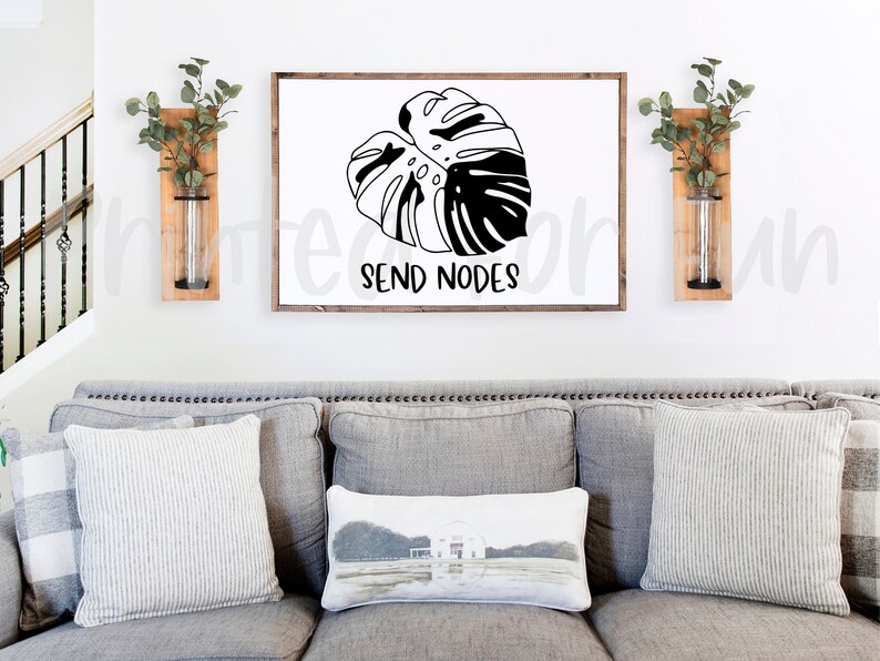May include: A white framed print with a black and white illustration of a monstera leaf. The text "SEND NODES" is below the leaf. The print is hanging on a white wall above a gray couch with pillows.