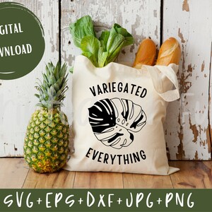May include: A white tote bag with black text that reads "Variegated Everything" and a black silhouette of a monstera leaf. The bag is sitting on a wooden surface with a pineapple and some bread in the background.