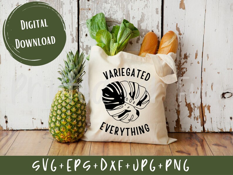 May include: A white tote bag with black text that reads "Variegated Everything" and a black silhouette of a monstera leaf. The bag is sitting on a wooden surface with a pineapple and some bread in the background.