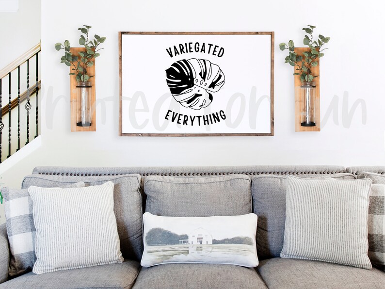 May include: A white framed sign with black text that reads "Variegated Everything" and a black silhouette of a monstera leaf. The sign is hanging on a white wall above a grey sofa with cushions. Two wooden wall planters with clear glass tubes are on either side of the sign.