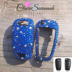 May include: Two blue and black car key covers with rhinestones. The covers are for a car key with two buttons. The key covers are on a wooden surface. The image also shows two black car keys with the Audi logo.
