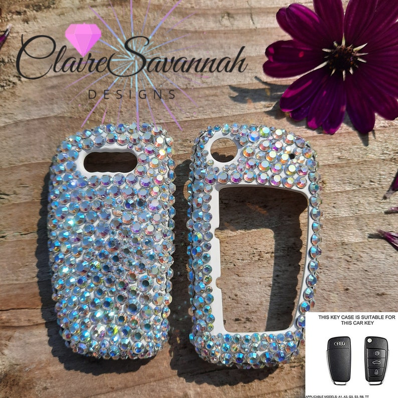 May include: Two white car key covers encrusted with iridescent rhinestones. The key covers are designed to fit Audi car keys. The text "THIS KEY CASE IS SUITABLE FOR THIS CAR KEY" is printed on a white background with a black Audi logo. Two black Audi car keys are shown below the text.