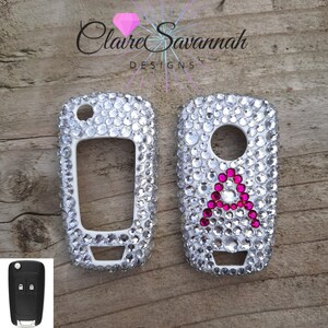 May include: Two white car key covers encrusted with clear rhinestones. One cover has a pink letter "A" in the center.  Claire Savannah Designs.