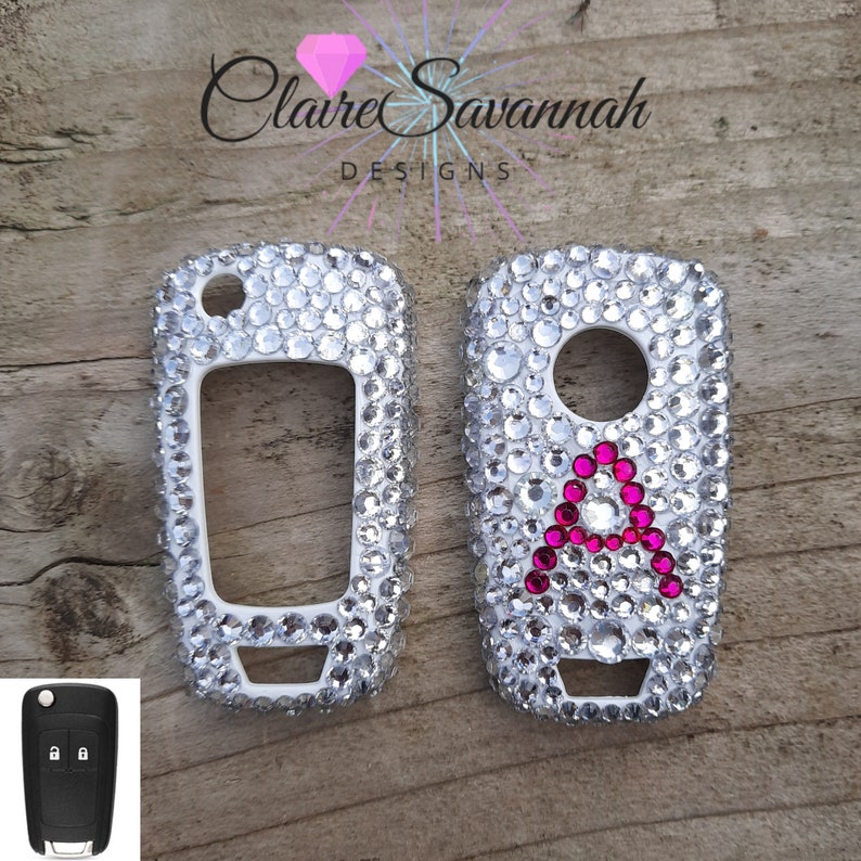 May include: Two white car key covers encrusted with clear rhinestones. One cover has a pink letter "A" in the center.  Claire Savannah Designs.