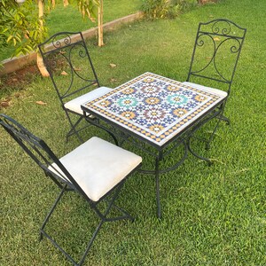 Puede incluir: Un juego de patio de metal negro con una mesa cuadrada y dos sillas. La superficie de la mesa está hecha de azulejos de mosaico coloridos con un patrón geométrico. Las sillas tienen cojines blancos.