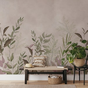 Op de afbeelding: Een interieur met een botanisch thema, met een bank met een geweven zitting, een strohoed en een decoratief kussen. De achtergrond is een muurschildering met groene en bruine bladeren tegen een gedempte roze achtergrond. Een potplant in een geweven mand staat op een zwarte stoel.