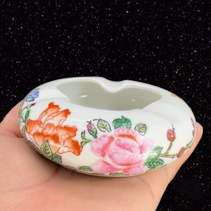 May include: White ceramic ashtray with a floral design. The design features pink, orange, and green flowers with gold accents.