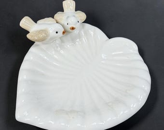May include: A white ceramic trinket dish shaped like a heart with a scalloped, shell-like texture. Two small, light beige and white birds with brown beaks are perched on the upper left edge of the dish, facing toward the center of the bowl.